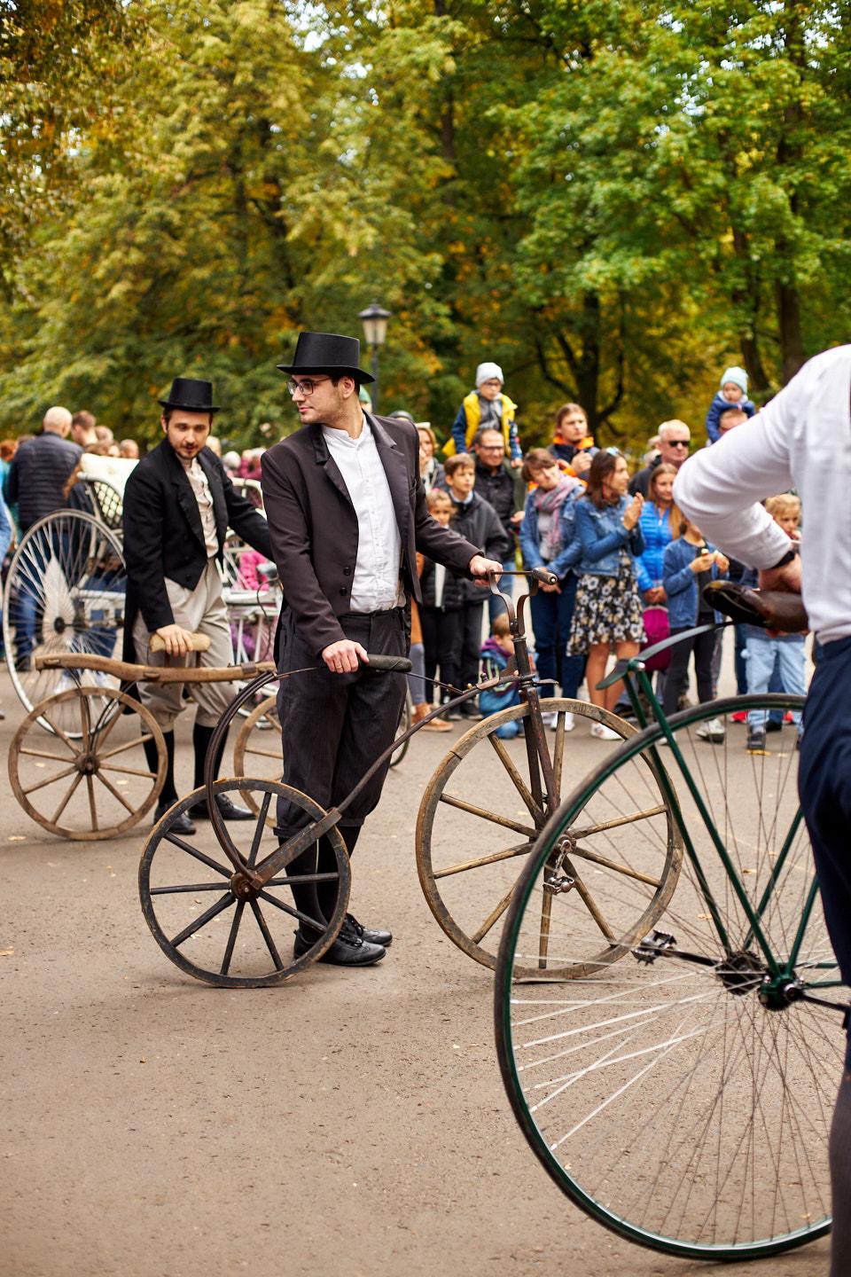 Przebrani w stroje z dwudziestolecia międzywojennego panowie stoją z bicyklami na asfaltowym placu. W tle drzewa Ogrodu Saskiego.