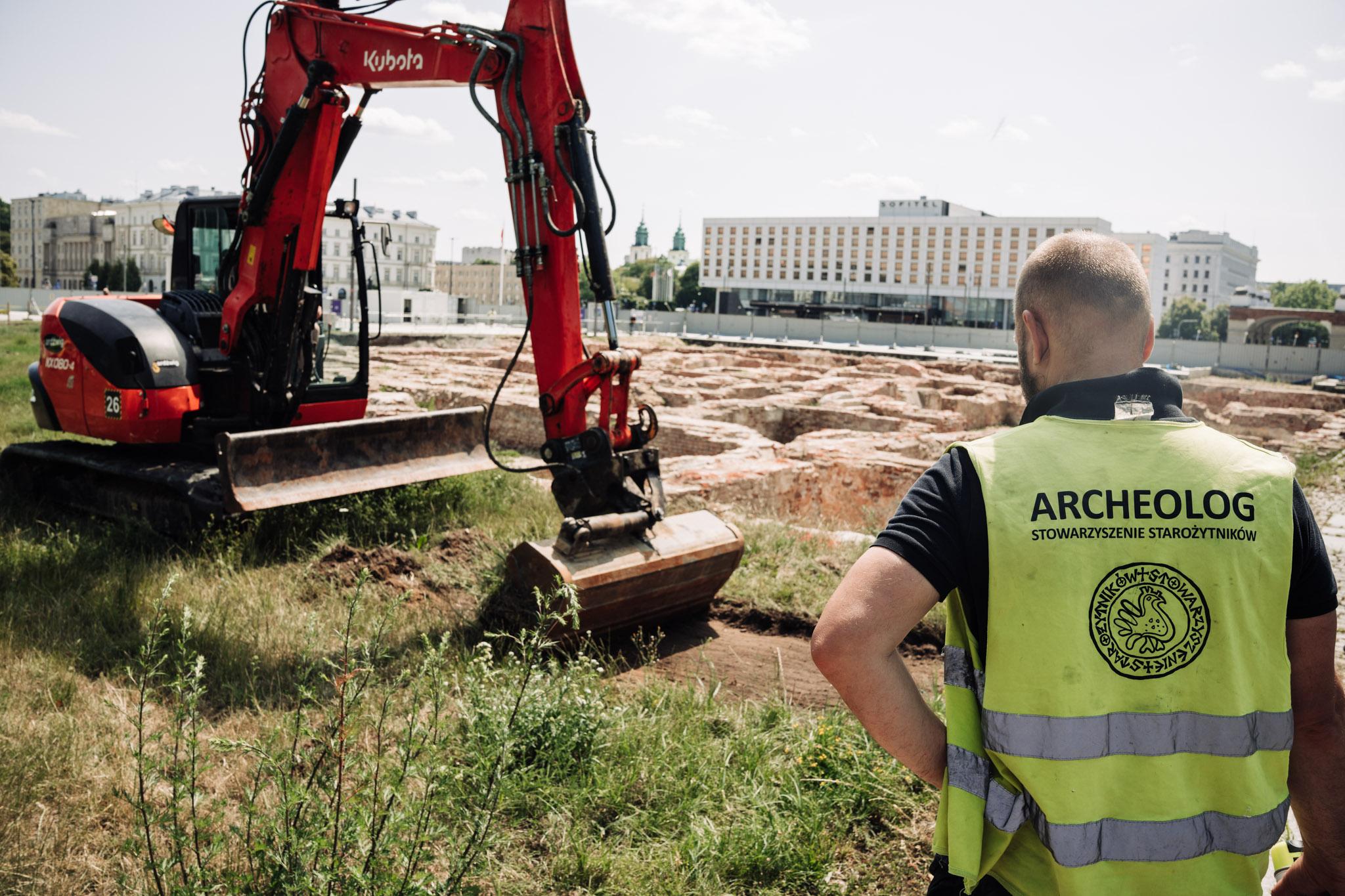 Na pierwszym planie archeolog w żółtej kamizelce z napisem "ARCHEOLOG STOWARZYSZENIE STAROŻYTNIKÓW" nadzorujący pracę koparki ściągającej z ziemi warstwę trawy. W tle odsłonięte już relikty Pałacu Saskiego.