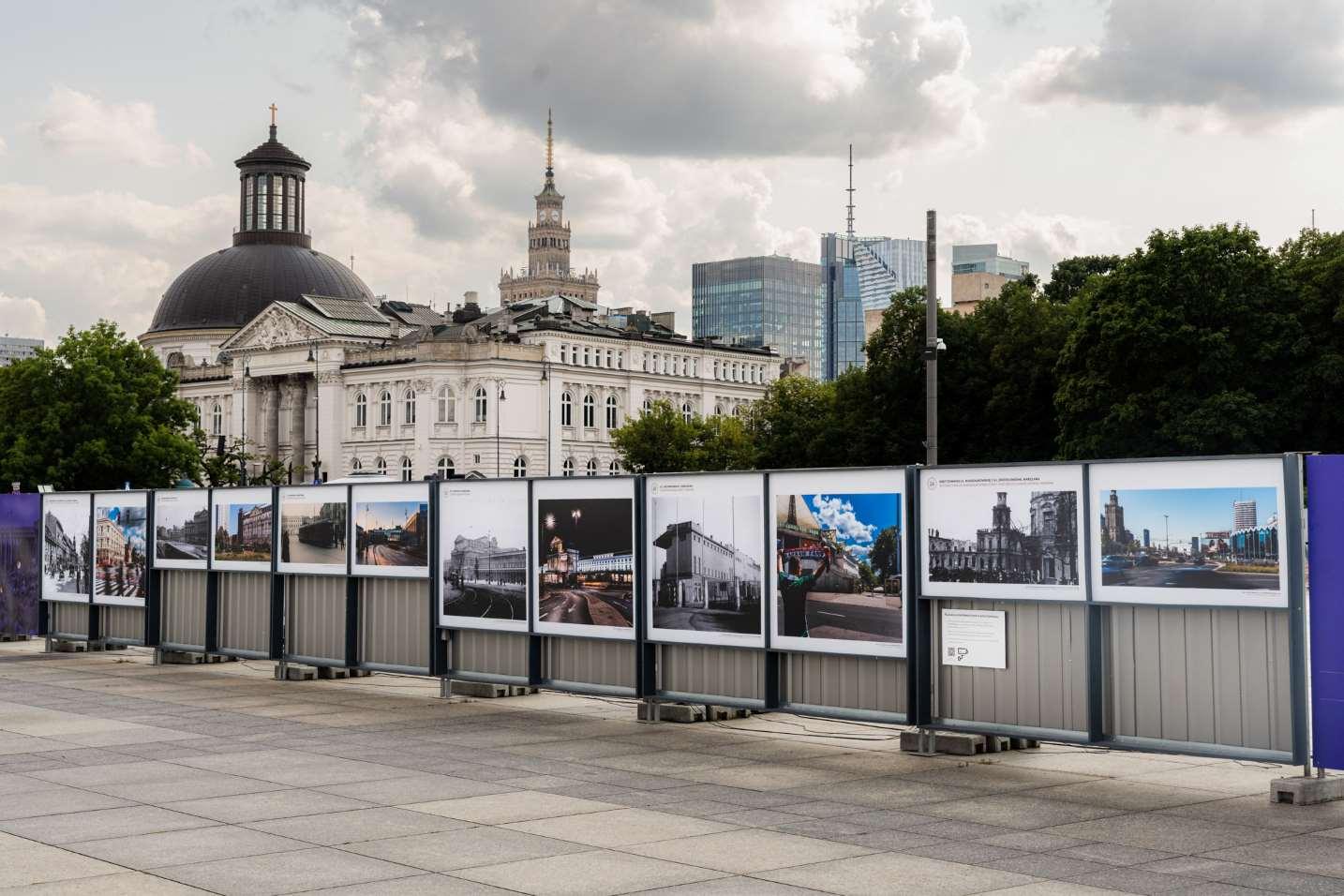 Widok z ukosa na wystawę zdjęciową na metalowym ogrodzeniu. W tle od lewej panorama Warszawy, po prawej drzewa Ogrodu Saskiego. Niebo częściowo zachmurzone.